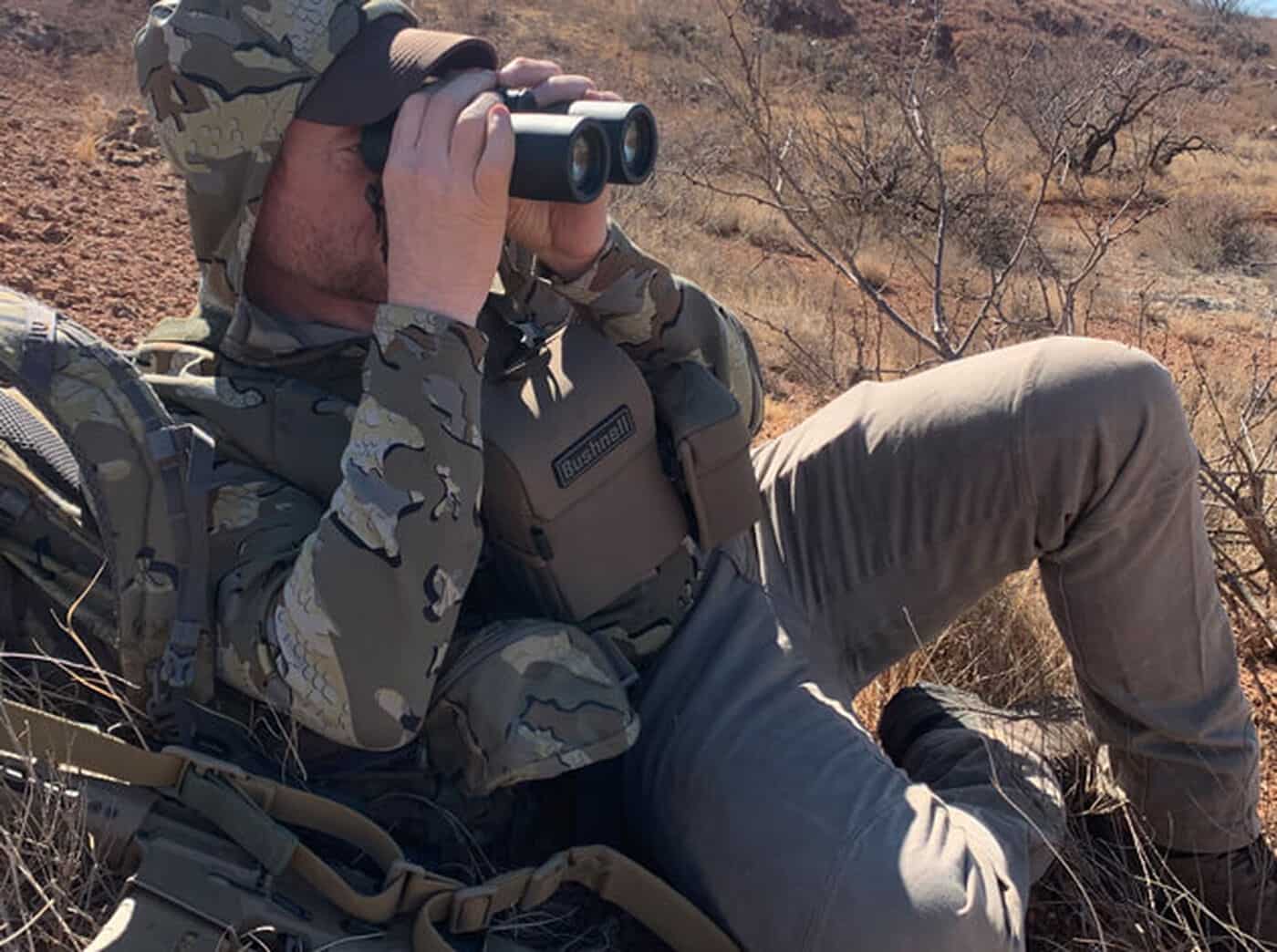 Man in wearing camo, in desert terrain, looking through binoculars while wearing Vault Binocular Pack